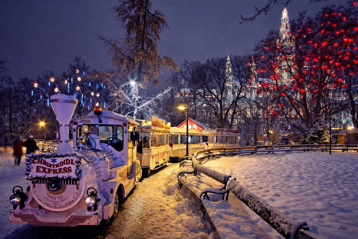Viyana'da kış mevsiminde karla kaplı bir parkta, önünde Christkindl Express yazan bembeyaz bir tur treni ilerlerken, arkadaki ağaçlar kırmızı kalpli ışıklarla süslü ve gotik mimarili büyük bir bina ışıklar içinde parlıyor.