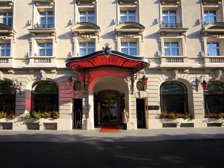 Valerie Messika'nın Paris Gezi Rehberi'nde önerdiği, kırmızı tenteli, neoklasik mimariye sahip The Royal Monceau Hotel Paris'in görkemli girişi, kapıda duran bir görevli, kırmızı halı ve pencerelerin altındaki çiçeklerle zarif bir atmosfer oluşturuyor.