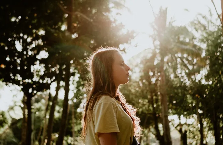 Güneş ışıkları altında doğada gözlerini kapatarak meditasyon yapan genç kadın.