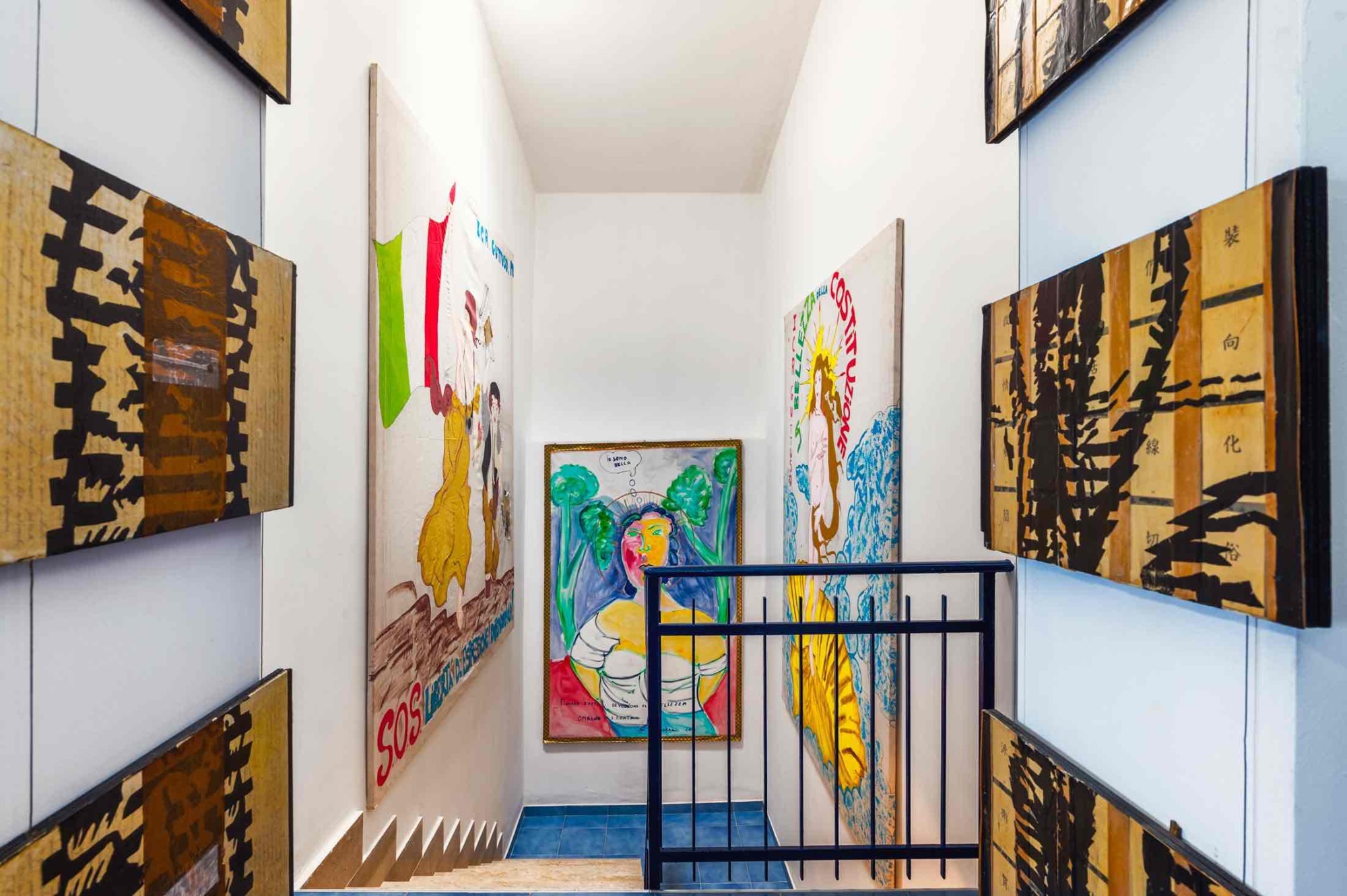 Atelier Sul Mare sanat otelinin dar merdiven boşluğu, beyaz duvarları süsleyen çeşitli sanat eserleriyle bir galeri gibi işlev görüyor. Mavi korkuluklar, canlı renkli tabloları ayırıyor.