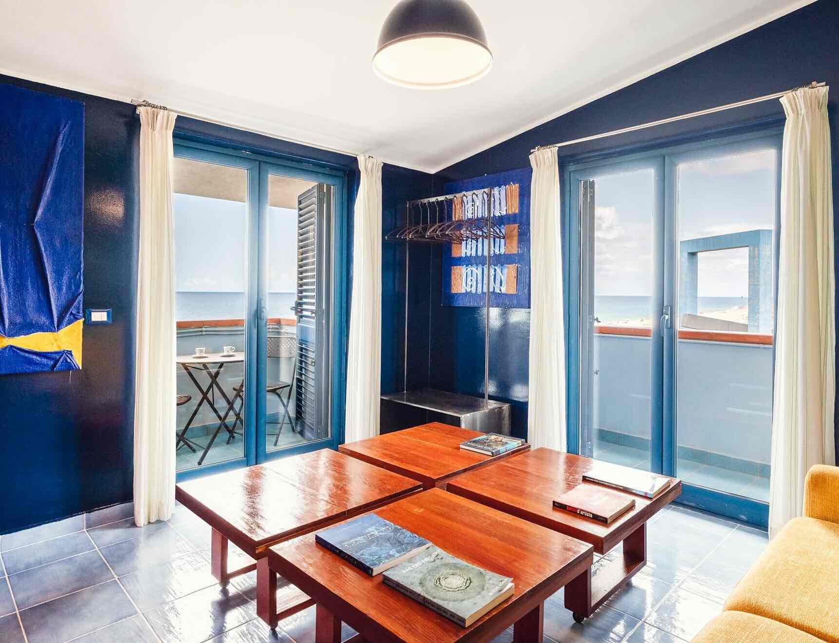 Atelier Sul Mare sanat otelinin deniz manzaralı odası. Koyu mavi duvarlar ve parlak ahşap sehpalar, balkondan sızan gün ışığıyla aydınlanıyor ve sanatsal bir atmosfer yaratıyor.