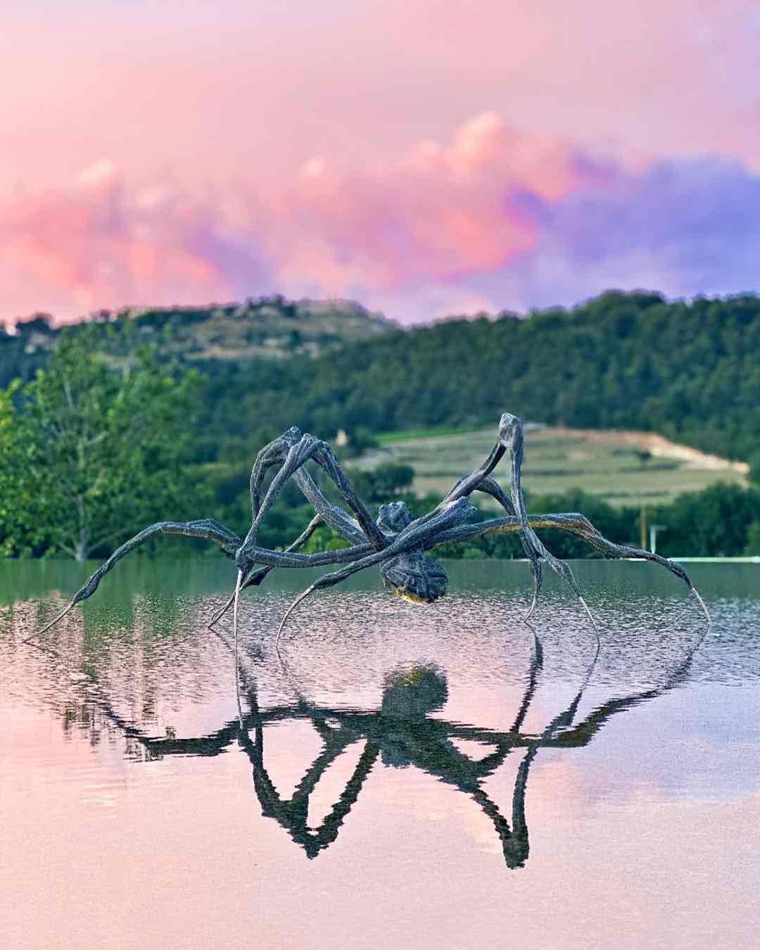 Château La Coste sanat parkındaki dev örümcek heykeli, sakin su yüzeyine yansıyor. Pembe gün batımı gökyüzü ve yeşil tepeler, esere dramatik bir fon oluşturarak sanat ve doğayı birleştiriyor.