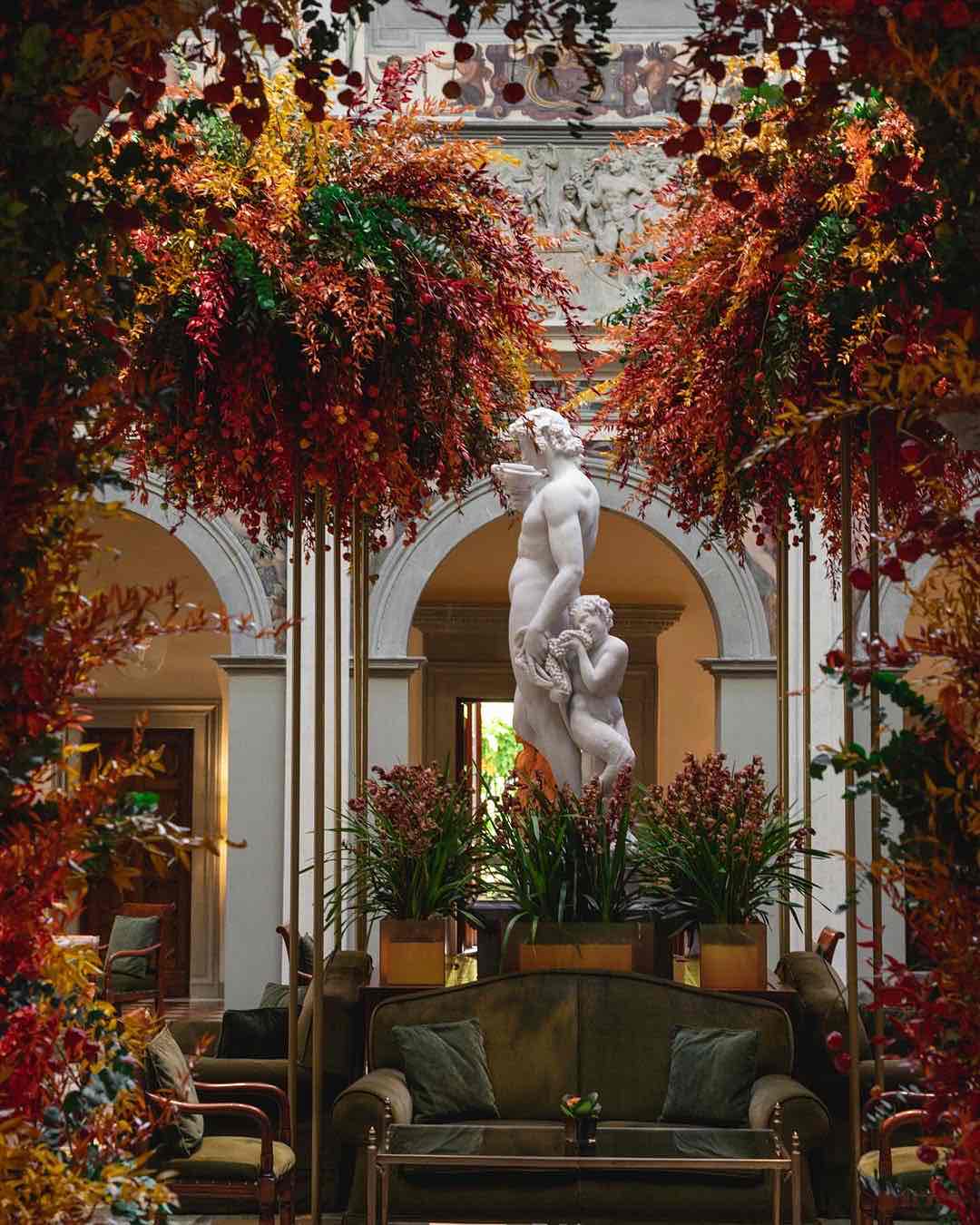 Four Seasons otelinin avlusunda, sonbahar renklerindeki devasa çiçek aranjmanları, kemerli geçitlerin önünde duran beyaz mermer heykeli sanatsal bir şekilde çerçeveliyor.