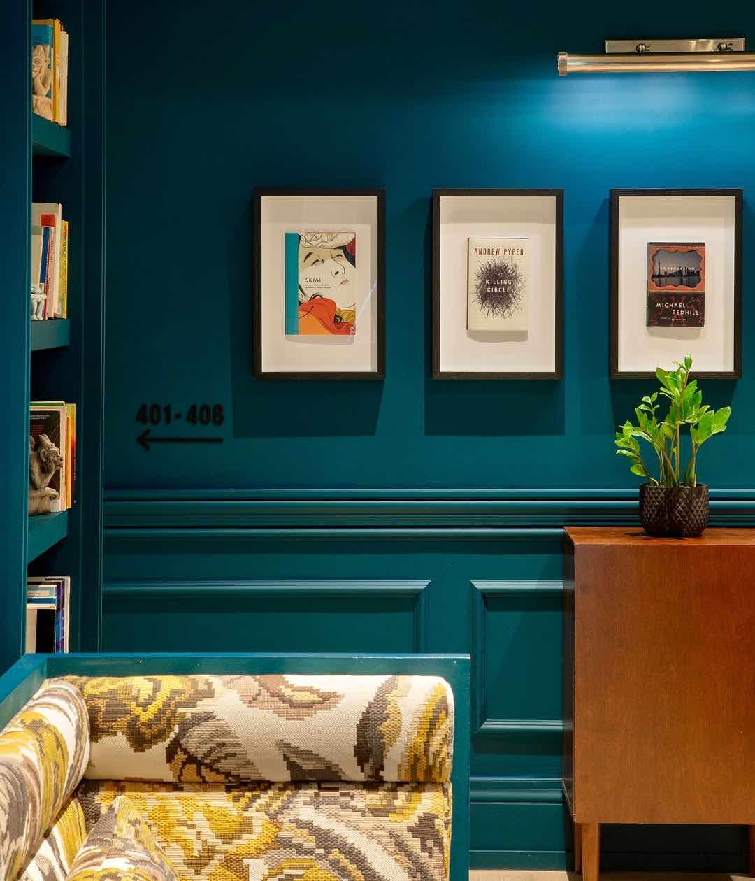 Gladstone Hotel'in edebi temalı iç dekorasyonu: Petrol mavisi duvarlar, çerçeveli kitap kapaklarını ve kitaplığı aydınlatırken, desenli koltuk ve ahşap konsol mekana sıcaklık katıyor.