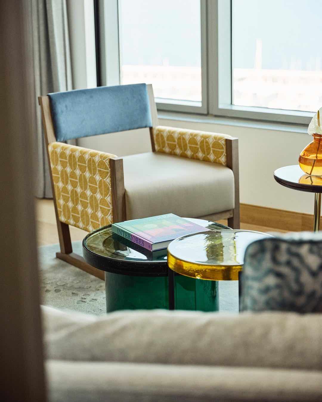 Hotel Arts süitinin pencere kenarında, mavi sırtlıklı ve sarı geometrik desenli kollara sahip tasarım koltuk. Yanında zümrüt yeşili ve sarı camdan modern sehpalar duruyor.