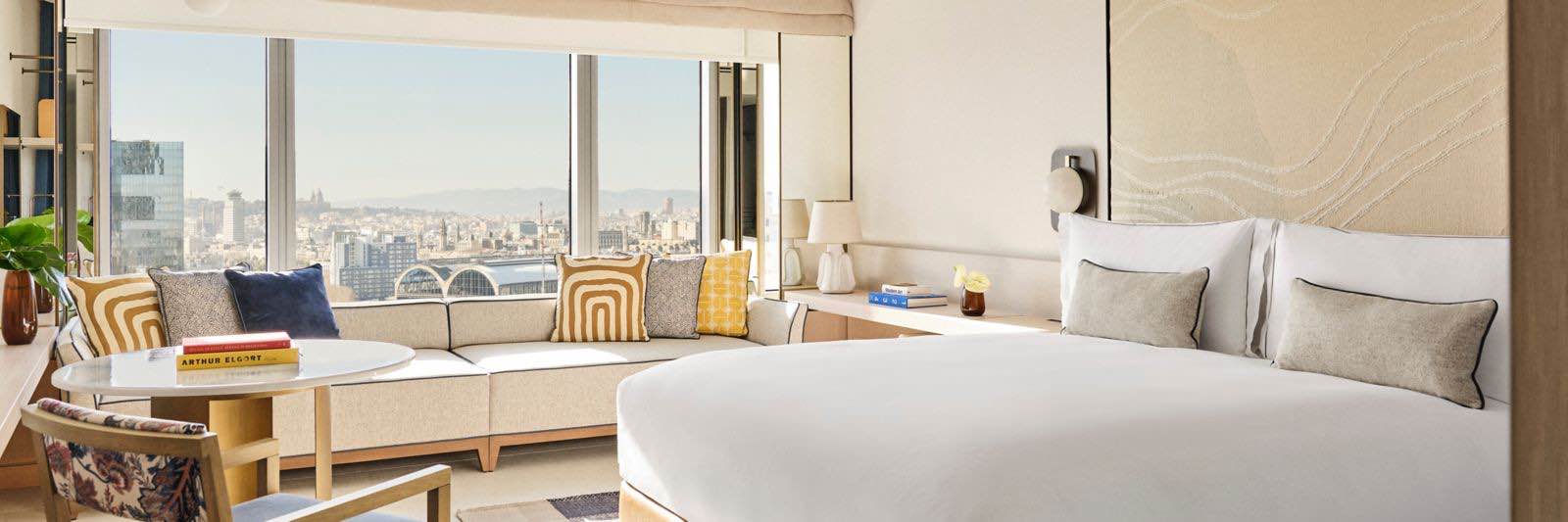 Hotel Arts'ın ferah ve aydınlık süiti, büyük pencerelerinden panoramik şehir manzarası sunuyor. Beyaz yatak takımları, bej kanepe ve renkli yastıklarla modern bir konfor alanı yaratılmıştır.