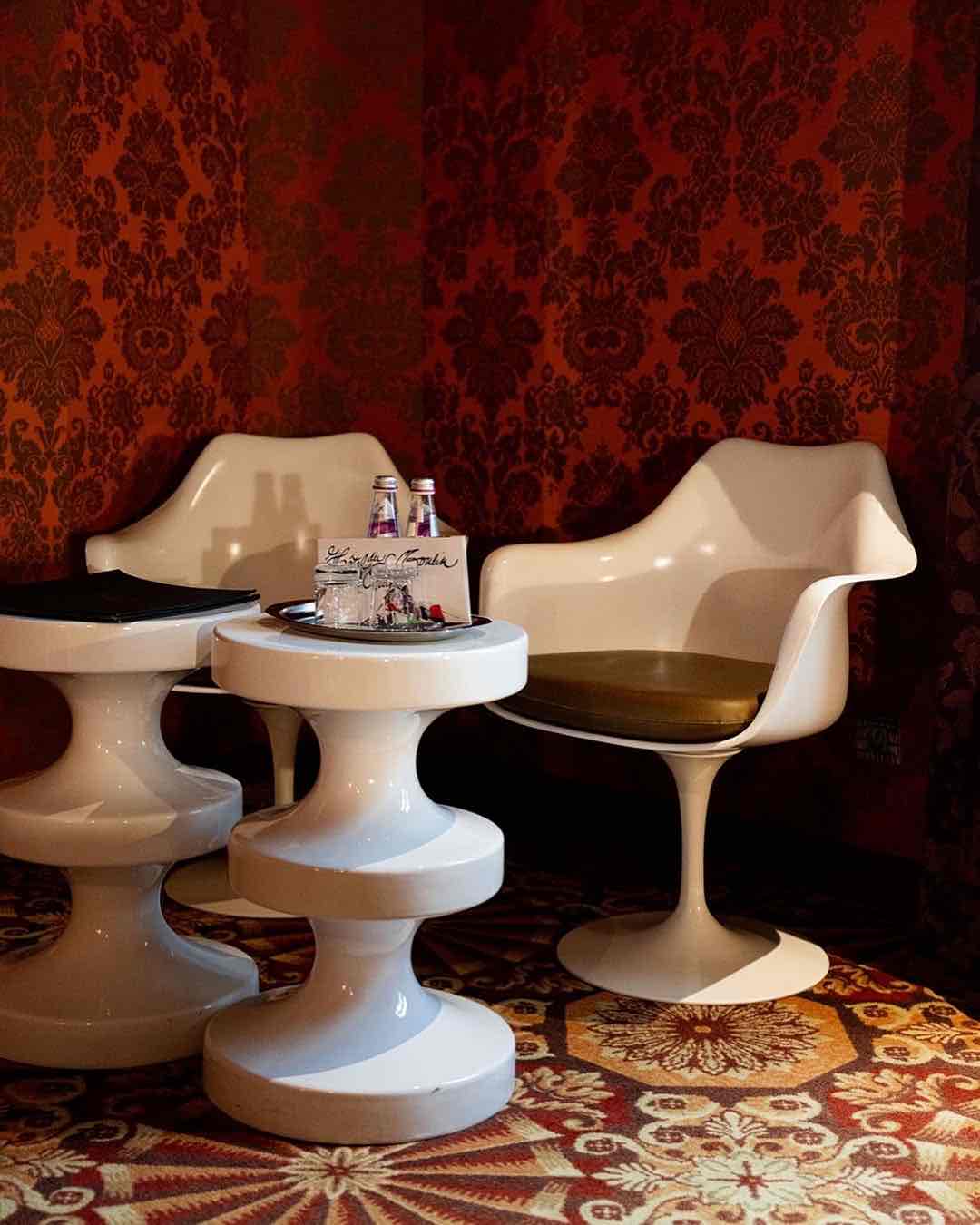 Hôtel du Petit Moulin'in eklektik tasarımını yansıtan bir köşe. Modern beyaz sandalyeler, zengin kırmızı damask duvar kağıdı ve desenli oryantal halı ile cesur bir kontrast yaratıyor.