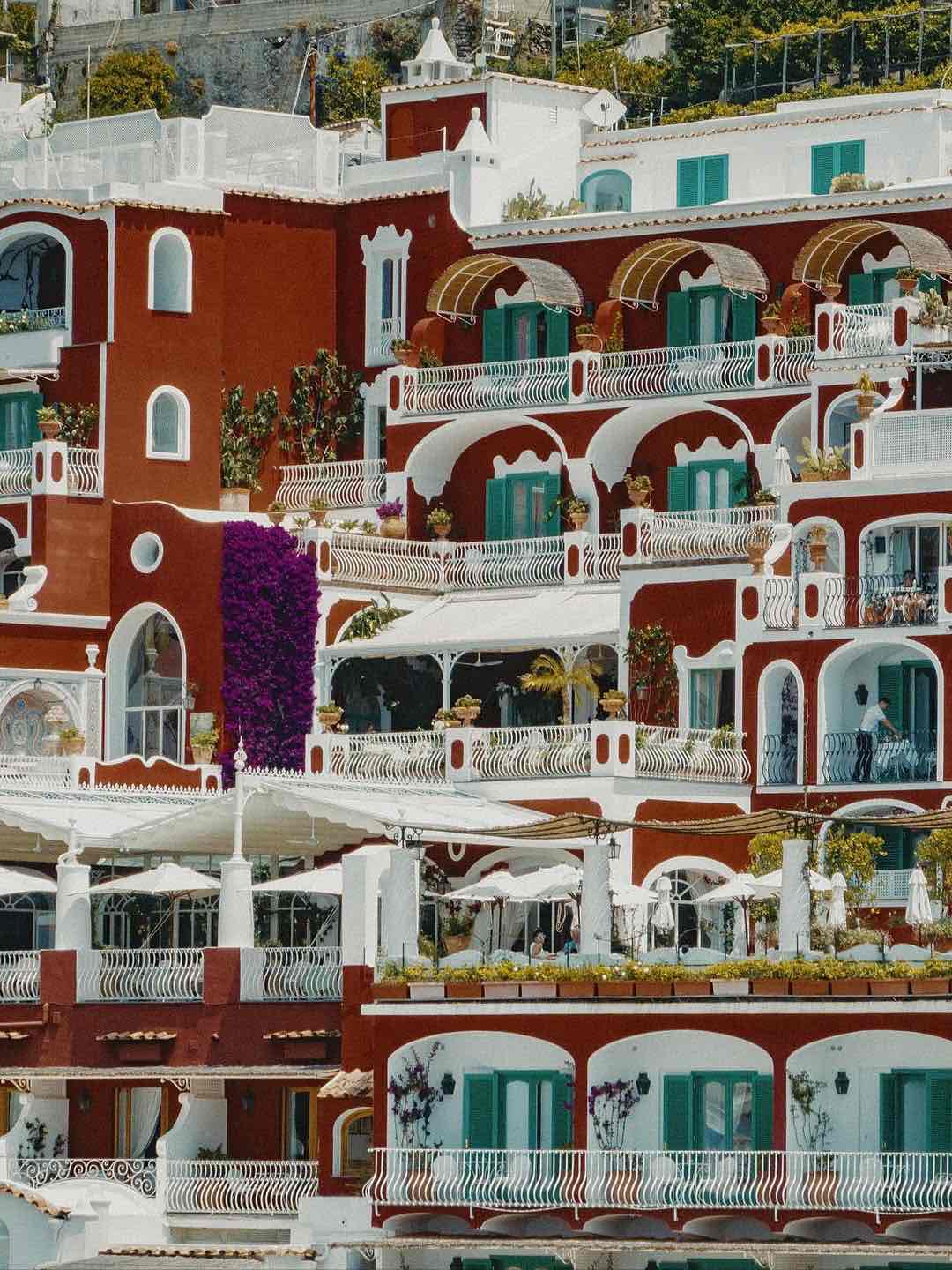 Positano'daki Le Sirenuse otelinin terakota kırmızısı cephesi. Beyaz kemerli balkonlar, yeşil panjurlar ve parlak mor begonviller, Amalfi Sahili'nin lüks ve katmanlı mimarisini gözler önüne seriyor.