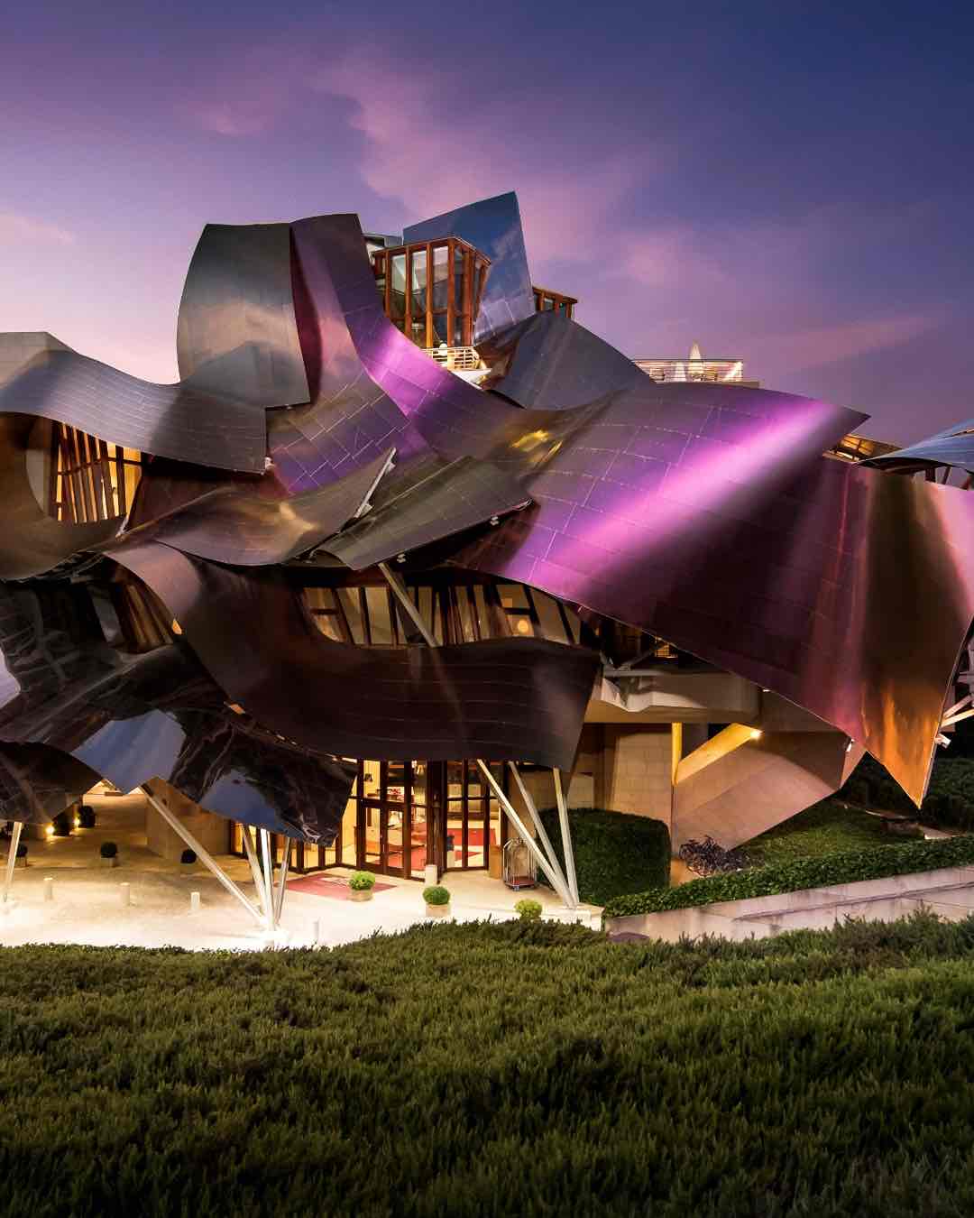 Hotel Marqués de Riscal'in heykelsi yapısı, gün batımında mor ve pembe ışıkları yansıtan dalgalı titanyum şeritlerle kaplı. Ön planda yeşil çalılar ve aydınlatılmış otel girişi yer alıyor.