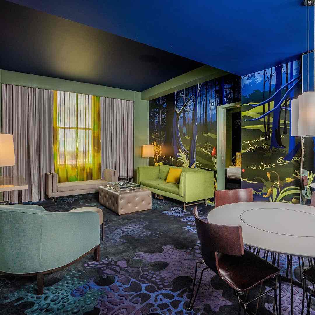 Bir Museum Hotel süitinde, duvarı kaplayan masalsı orman resmi ve koyu mavi tavan, sürükleyici bir atmosfer yaratıyor. Modern mobilyalar, mor ve mavi desenli halı üzerinde sanat ve konforu