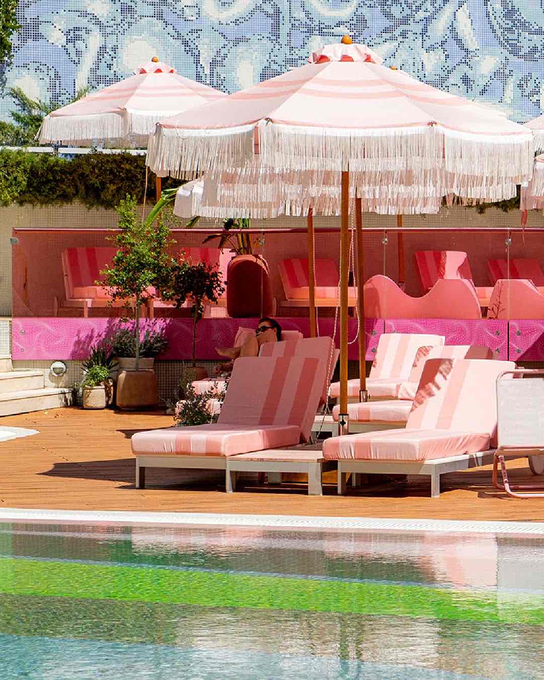 Semiramis Hotel'in canlı tasarımını yansıtan havuz alanı. Püsküllü pembe şemsiyeler, çizgili şezlongları gölgelendirirken, arkadaki mavi mozaik duvar sanatsal bir atmosfer yaratıyor.