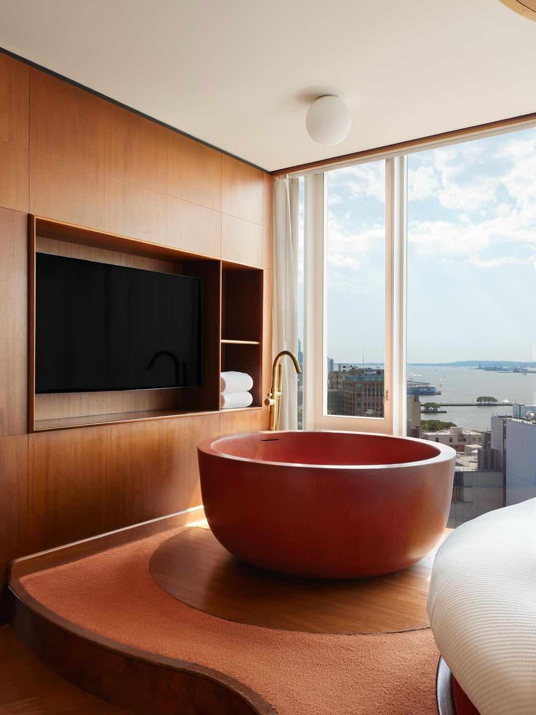 The Standard otel odasında, şehir ve su manzaralı büyük pencerenin önünde duran yuvarlak, kırmızımsı kahverengi küvet. Yatak odasının bir parçası olan küvet, ahşap panelli duvardaki TV'nin yanında
