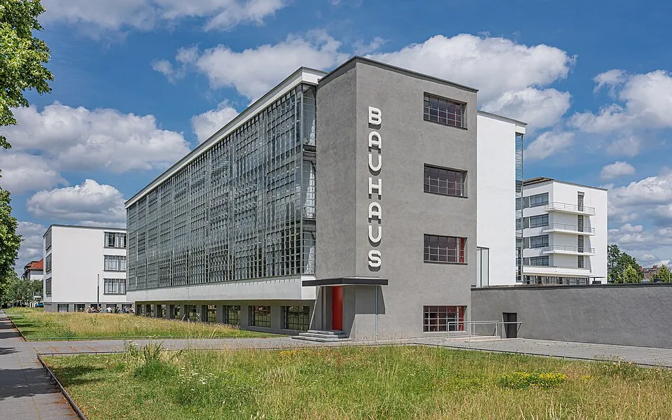 Dessau'daki ünlü Bauhaus Okulu binası, modern mimarinin karakteristik özelliklerini taşıyan gri ana bloğu, geniş cam cephesi ve kırmızı giriş kapısının üzerinde dikey olarak yazılmış büyük beyaz BAUHAUS yazısıyla masmavi gökyüzü altında göz alıcı duruyor.