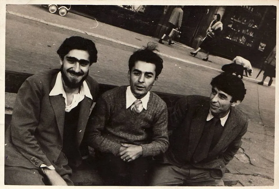 Siyah beyaz fotoğrafta, kaldırım kenarındaki bir bankta yan yana oturan genç üç erkek görülüyor; sol baştaki sakallı Sadi Öziş genişçe gülümserken, ortadaki örgülü kazak ve kravatıyla İlhan Koman dikkatle bakıyor, sağdaki arkadaşları ise onlarla sanat ve tasarım üzerine konuşurken hafifçe gülümsüyor.