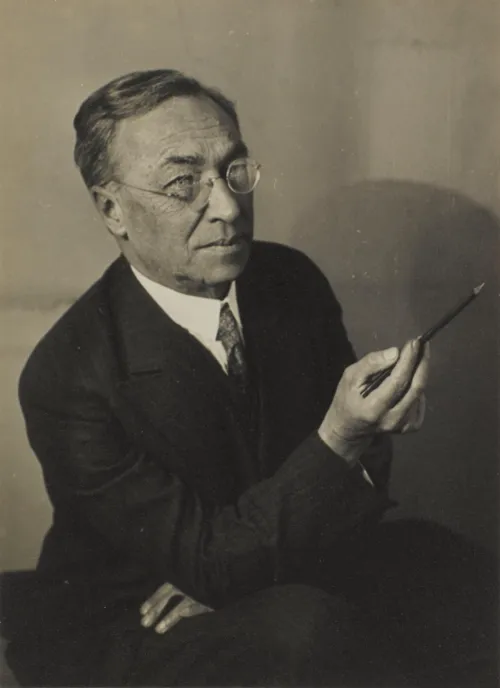 Siyah beyaz bu sanatsal portre fotoğrafında, ünlü ressam Vassily Kandinsky, düşünceli bir ifadeyle gözlüğü ve şık takım elbisesiyle kameradan hafifçe sağa doğru bakarken sağ elinde ince bir fırça tutuyor, onun yaratıcı ruhunu ve sanatla olan derin bağını vurguluyor.