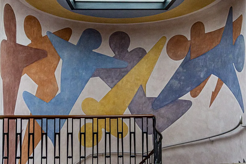 Weimar Bauhaus sanatının dinamik ruhunu yansıtan, eğri bir merdiven boşluğunun duvarını kaplayan, soyut insan figürleri ve sarı geometrik şekillerle kahverengi, turuncu, mavi ve mor tonlarında hareketli bir duvar resmi, üstte bir ışıklık ve altta siyah metal bir korkulukla görülüyor.