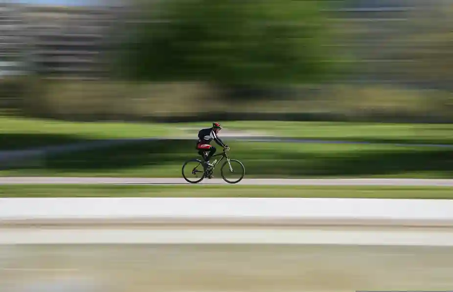 Kask ve sırt çantası takmış, özel giyimli bir bisikletçi, hızın etkisiyle bulanıklaşmış yemyeşil bir doğa ortamında, pürüzsüz bir bisiklet yolunda pedal çevirerek, en iyi bisiklet rotalarından birinde özgürce ilerlemenin keyfini çıkarıyor.