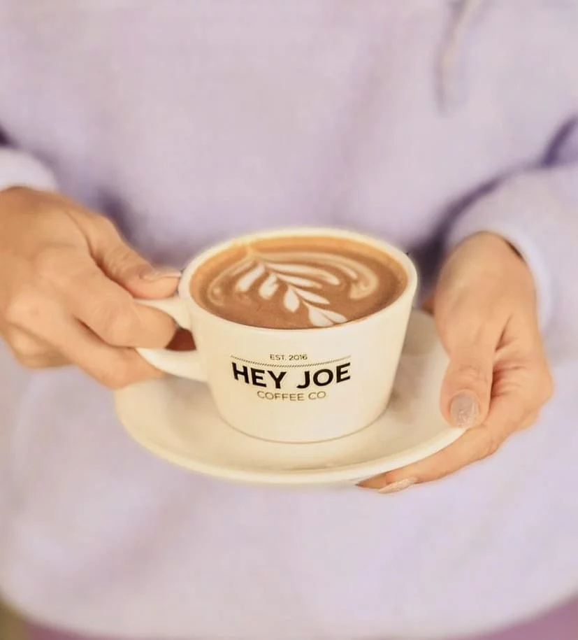 Açık mor renkli bir kazak giyen bir kişinin elleri, estetik yaprak deseniyle süslenmiş bol köpüklü, üzerinde EST. 2016 HEY JOE COFFEE CO logosu olan beyaz bir fincan latteyi altlığıyla birlikte nazikçe tutuyor.