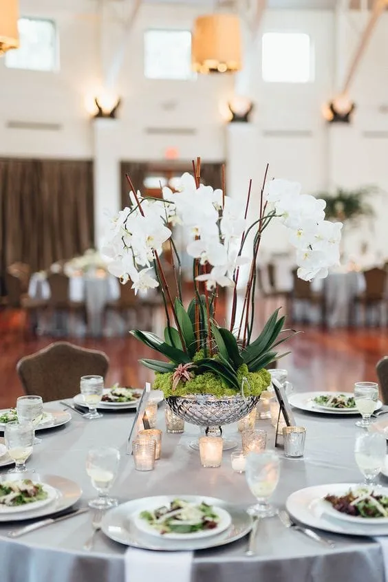 Gümüş detaylı büyük bir kasede sergilenen beyaz Phalaenopsis orkideleri, yeşil yosun ve geniş yapraklarla zenginleştirilmiş. Şık bir düğün veya davet masasının tam ortasında konumlandırılan bu aranjman, etrafındaki mum ışıkları ve tabak düzeniyle romantik bir atmosfer yaratıyor.