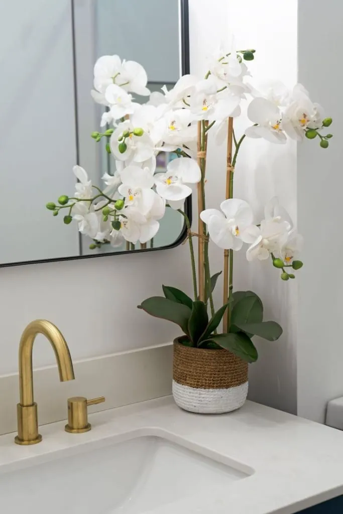 Modern bir banyoda, beyaz lavabonun yanında yer alan jüt kaplamalı saksıya dikilmiş beyaz Phalaenopsis orkideleri. Altın renkli musluk detayı ve sade tasarımla birlikte spa hissi uyandıran huzurlu bir köşe yaratıyor.