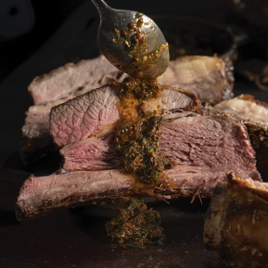 Ortadan az pişmiş, sulu görünümlü ızgara dana etinin üzerine bir kaşıkla Arjantin'in meşhur chimichurri sosunun döküldüğü bu yakın çekim kare, Buenos Aires'in zengin gastronomi kültürünü ve tutku dolu lezzetlerini çağrıştırıyor.