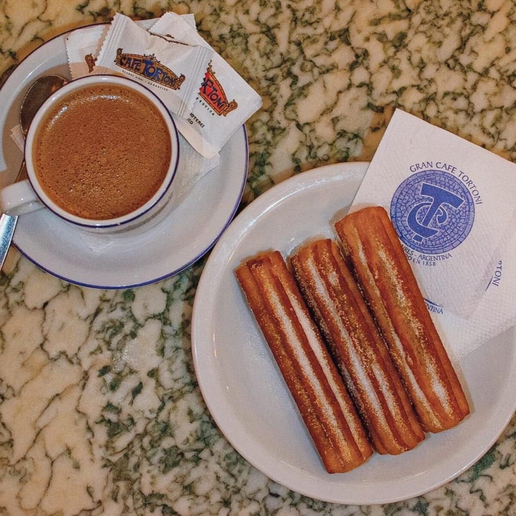Bir fincan kahve ve bol şekerli üç adet çıtır churros'un, Buenos Aires'in köklü kültürü ve tutkusunu yansıtan ikonik Cafe Tortoni logosunu taşıyan peçete ve şeker paketleriyle birlikte yeşil damarlı mermer bir masada duruşu, şehrin tarihi kafelerinde zamanın durduğu anları anımsatıyor.