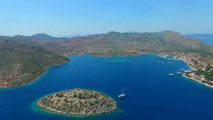 Bozburun'un masmavi sularla çevrili, yeşilimsi tepelerle sarılmış, berrak gökyüzü altında yelkenli teknelerin ve küçük bir adanın süslediği doğal limanı, kıyıdaki şirin kasaba evleriyle bütünleşen kuşbakışı görünümü.