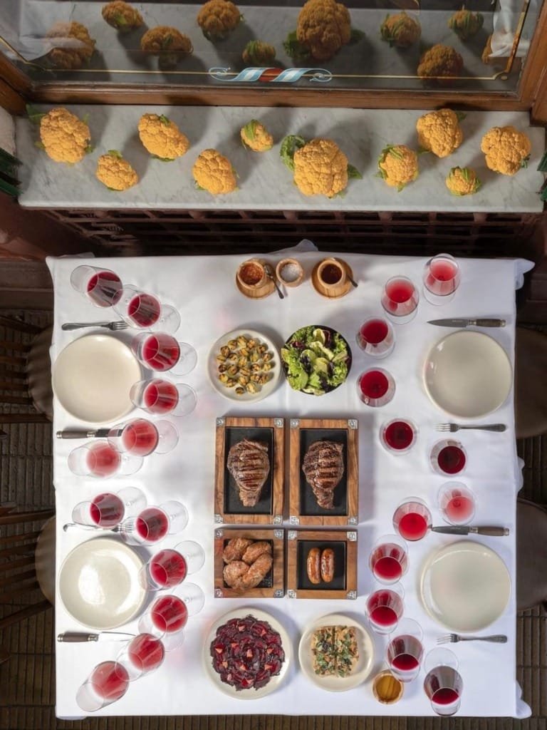 Beyaz masa örtülü geniş bir yemek masası, iki büyük ızgara biftek, yanında sosis, çeşitli mezeler ve bolca kırmızı şarap kadehleri ile Buenos Aires'in eşsiz gastronomi tutkusunu yansıtan, zengin bir Arjantin parrilla deneyimi sunuyor.