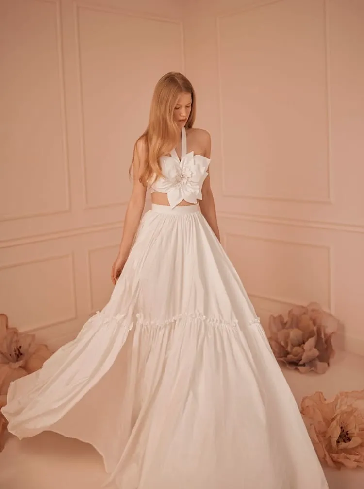 Pembe bir odada poz veren model, dev çiçek detaylı halter yaka crop top ve geniş, katmanlı etekten oluşan iki parçalı sade ve romantik Yael gelinlik giyiyor.