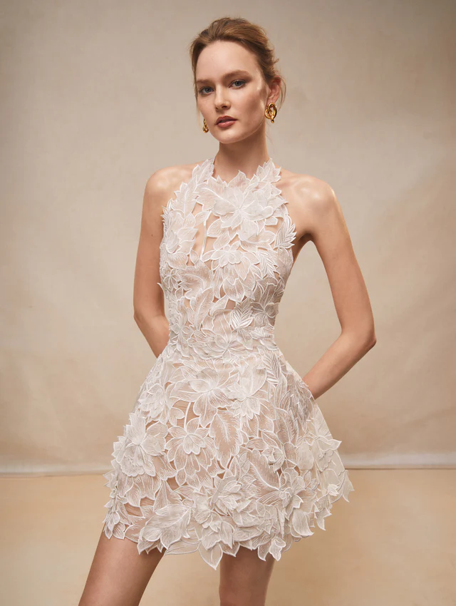 Oscar de la Renta 2026 koleksiyonundan, halter yaka detaylı ve 3D çiçek aplikeli kısa gelinlik modeli.