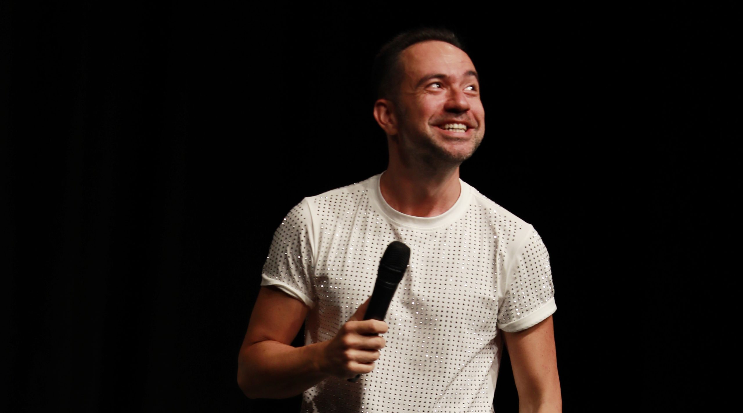 Sahnedeki Kaan Sekban, mikrofonuyla pırıltılı beyaz tişörtü içinde neşeyle yukarı bakarak, İstanbul etkinlik rehberi kapsamında şubat ayı stand-up gösterileriyle izleyicilere kurumsal hayattan ve sosyal medya gözlemlerinden beslenen mizahını sunuyor.