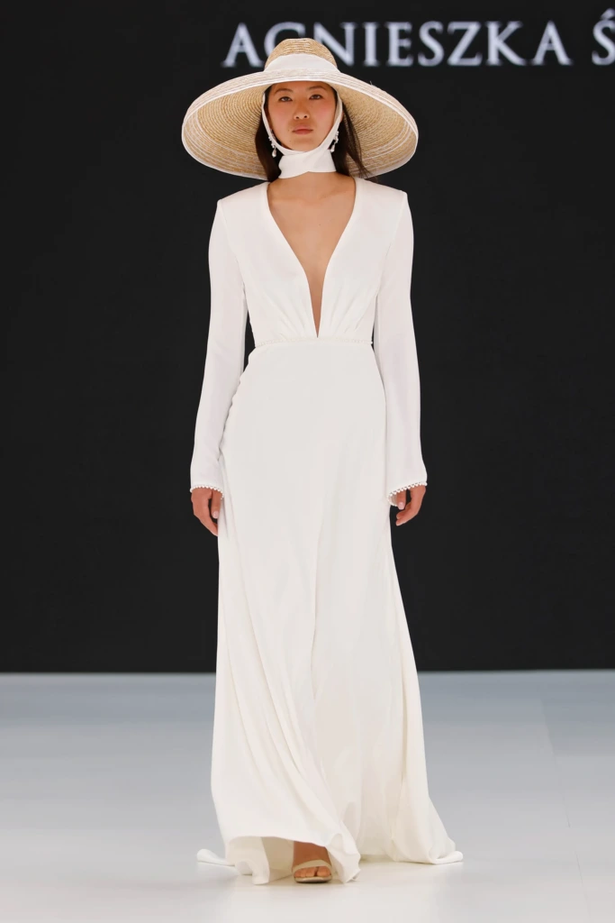 Derin V yaka kesimli, uzun kollu, sade ve akıcı kumaştan tasarlanmış, bel kısmında drape ve inci detaylı Agnieszka Światły marka modern minimalist gelinlik modeli, geniş hasır şapka ile tamamlanmış.