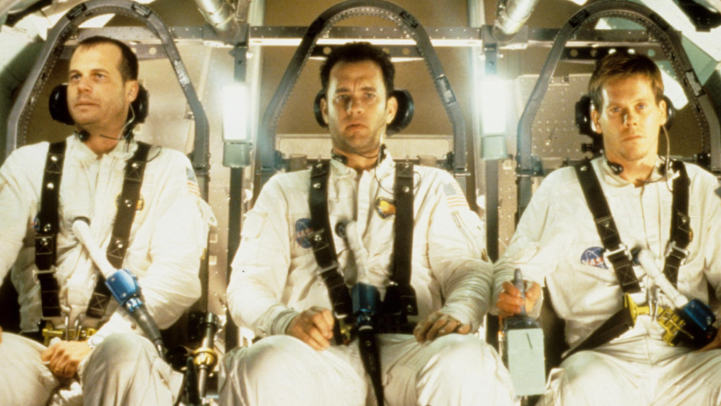 Apollo 13 filminden bir sahne: Üç astronot uzay aracının içinde yan yana otururken ciddi ifadelerle ileriye bakıyor. Üzerlerinde NASA amblemli beyaz astronot tulumları var ve kemerlerle sabitlenmiş durumdalar. Ortam, uzay görevlerinin gerilimli atmosferini yansıtıyor.
