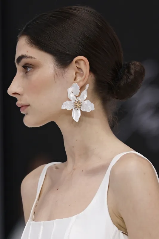 Badgley Mischka defilesinden çarpıcı bir görünüm: Kristal detaylı büyük beyaz çiçek küpeler ve sade kesimli zarif gelinlik ile romantik ve cesur bir stil birleşimi.