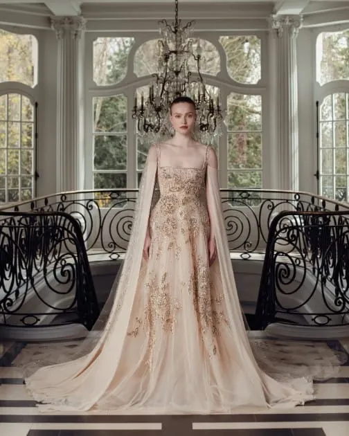 Georges Hobeika tasarımı, altın işlemeli nude tonda A kesim gelinlik modeli. İnce askılı ve pelerin detaylı bu gösterişli tasarım, zarif ışıltısı ve ihtişamlı duruşuyla saray atmosferinde büyüleyici bir görünüm sunuyor.