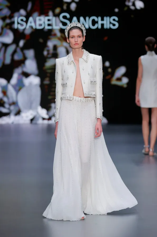 Isabel Sanchis 2025 İlkbahar koleksiyonundan, taş işlemeli kısa ceket ve yüksek bel transparan etekle tamamlanan modern bridal takım. Kraliyet tacı aksesuarıyla güç ve zarafeti buluşturan alternatif gelinlik stili.