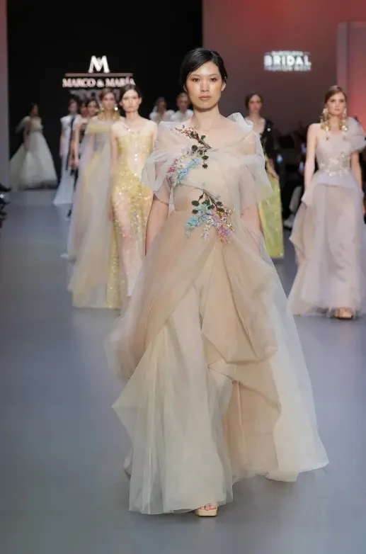 Marco & Maria 2025 İlkbahar koleksiyonundan, ten rengi tül katmanlarla tasarlanmış ve renkli çiçek aplikeleriyle süslenmiş transparan gelinlik modeli. Yüksek moda dokunuşlarıyla romantik ve bohem çizgileri buluşturan sanatsal bir görünüm.