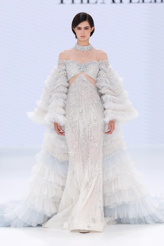 The Atelier by Jimmy Choo tasarımı, ışıltılı işlemelerle bezenmiş, off-shoulder kesimli, transparan bant detaylı ve dramatik tül kollu gösterişli gelinlik modeli.