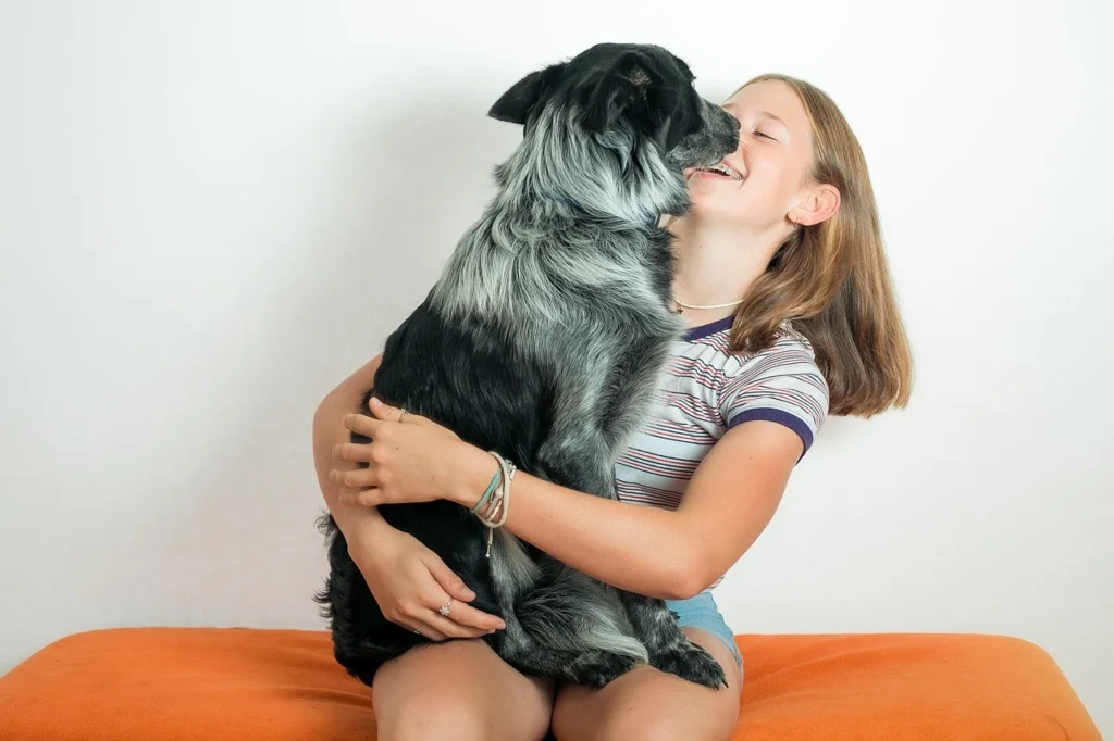 Gülümseyen genç bir kız, kucağında oturan büyük siyah-gri tüylü köpeğini sarılarak severken yakın bir bağ kuruyor.