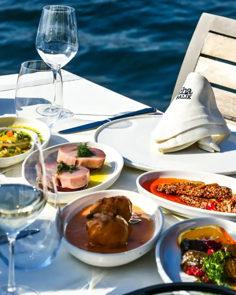 Deniz kenarında güneşli bir havada, Cha Cha Balık restoranının beyaz masasında, çeşitli taze balık mezeleri, zeytinyağlılar ve Akdeniz lezzetleri, zarif kadehler ve markalı peçete eşliğinde göz alıcı bir sunumla yer alıyor.