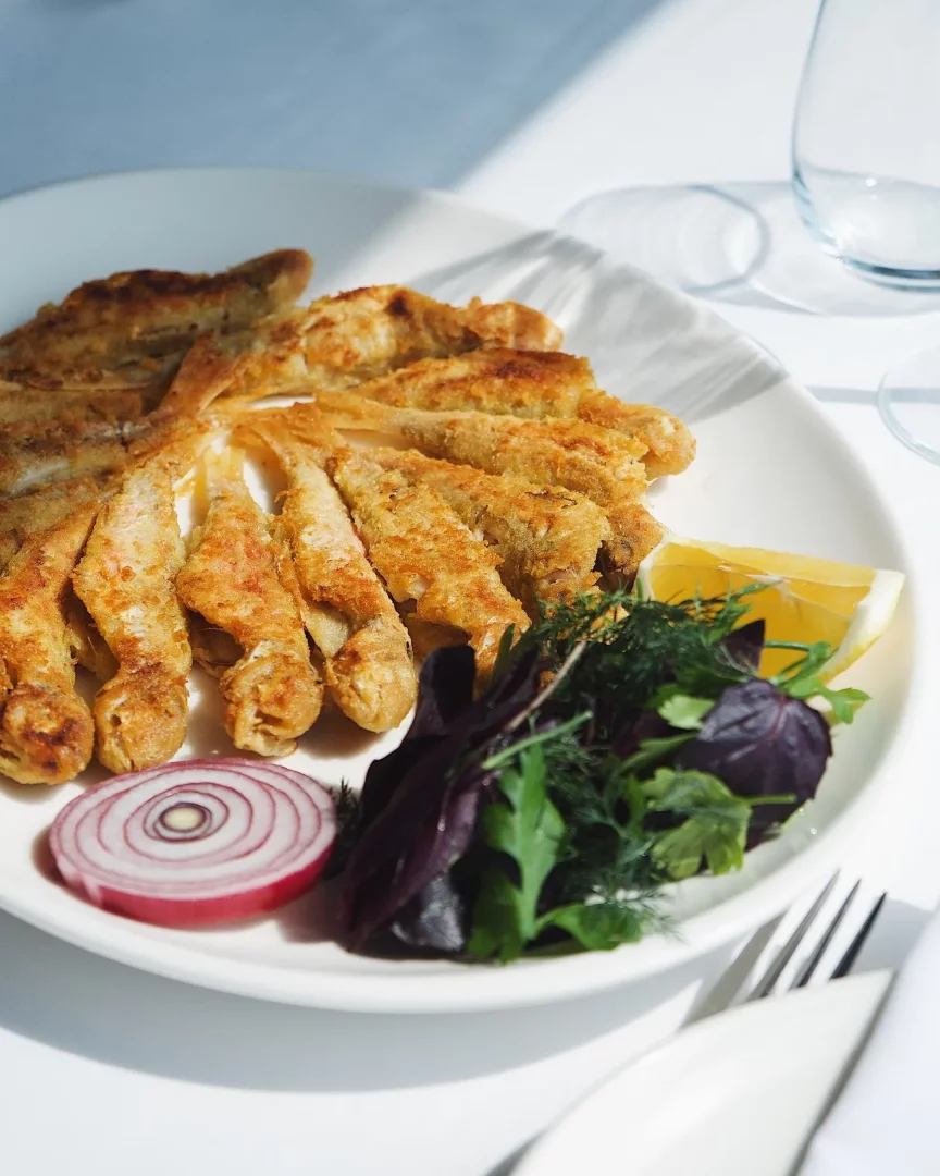 Beyaz bir tabakta güneş ışıkları altında servis edilmiş, çıtır çıtır kızarmış küçük balıklar, yanında taze yeşillikler, mor soğan dilimi ve limon ile iştah açıcı bir deniz mahsulü yemeği.