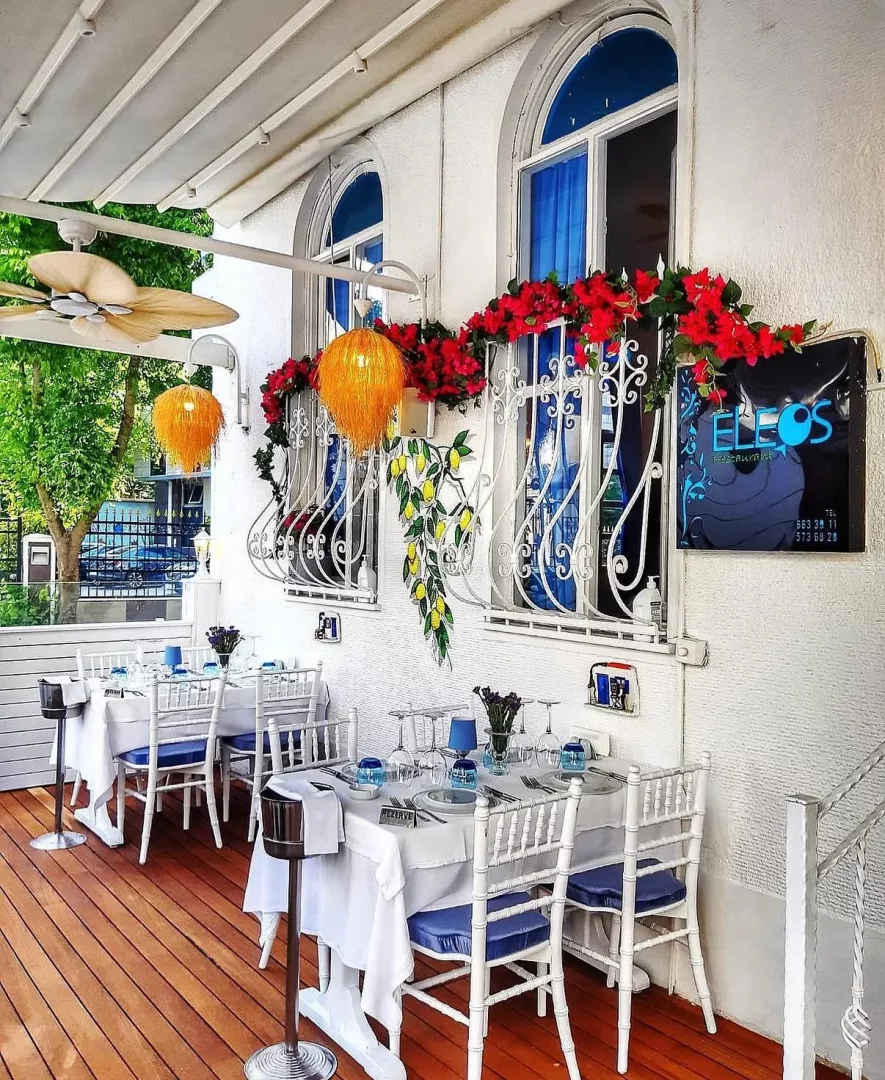 Eleos Restaurant'ın ahşap zeminli, beyaz tavan perdesi altında yer alan şık açık hava terası; beyaz masa örtülü, mavi minderli sandalyelerle düzenlenmiş masaları, kemerli pencereleri çevreleyen kırmızı çiçekleri, limon motifli süslemeleri ve turuncu, tüylü avizeleriyle göz alıcı bir atmosfer sunuyor.
