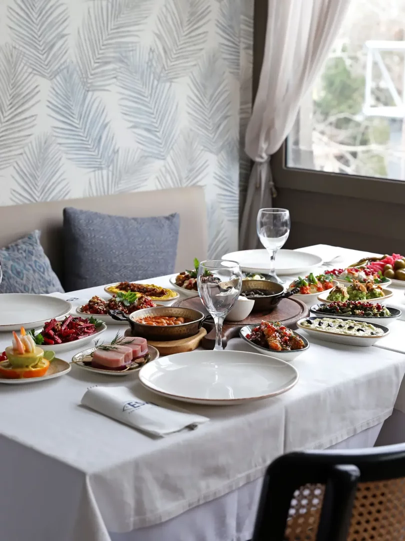Palmiye yapraklı duvar kağıdı ve dışarıyı gösteren pencere önünde, zeytin, humus, pancar salatası, kuru fasulye ve karides kokteyli gibi birçok geleneksel meze tabağıyla dolu bir Türk sofrası, eski yer atmosferini yansıtan sıcak bir restoranda davetkar bir şekilde sergileniyor.