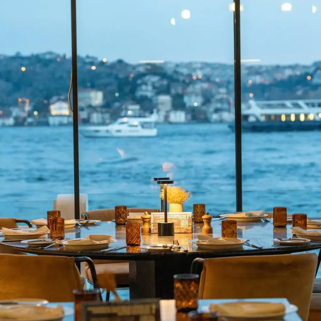 Boğaz manzaralı bir restoranda, şık bir masa düzeniyle akşam yemeği için hazırlanmış yuvarlak masa, suya yansıyan şehir ışıkları ve yakından geçen tekneler eşliğinde davetkar bir atmosfer sunuyor.