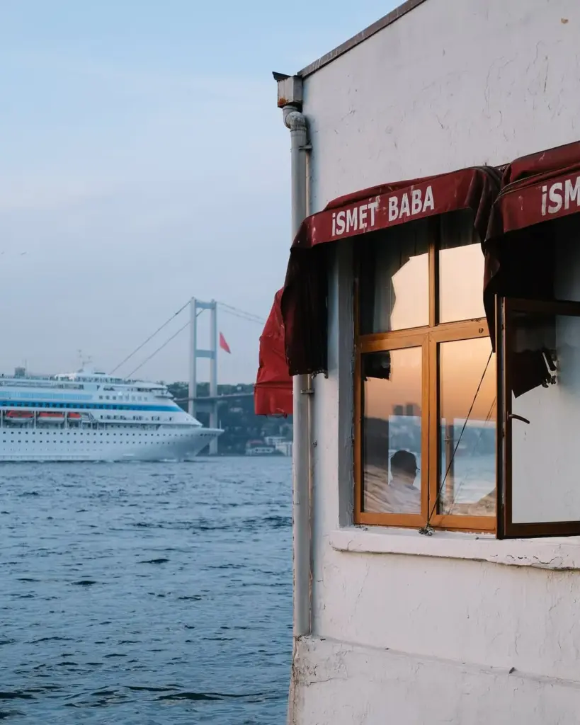İsmet Baba restoranının ahşap pencerelerinden yansıyan gün batımı kızıllığı, Boğaz'ın serin sularında ilerleyen büyük bir kruvaziyer gemisi ve arka plandaki köprü manzarasıyla, eşsiz bir İstanbul ambiyansını gözler önüne seriyor.