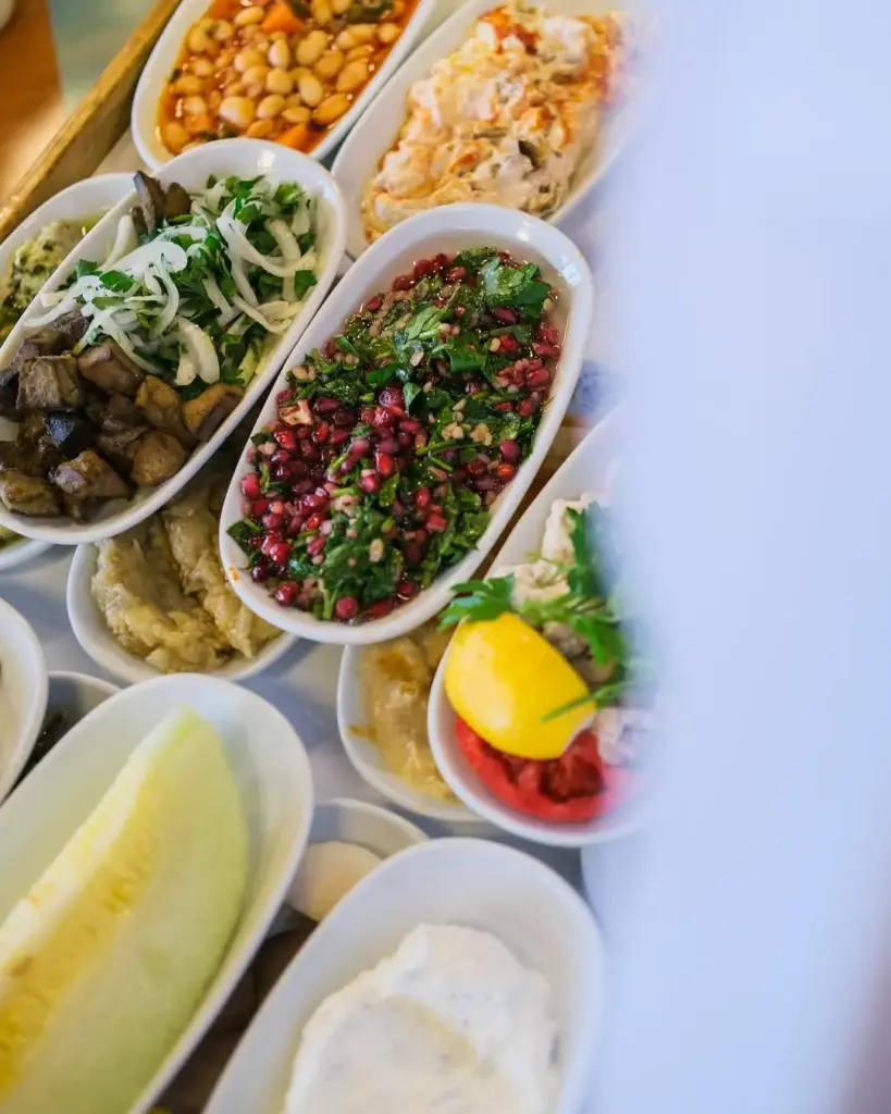 İsmet Baba restoranında sunulan zengin meze çeşitlerinden oluşan masa üst görünümü; beyaz oval tabaklarda barbunya pilaki, közlenmiş patlıcanlı salata, nar taneli yeşil salata, limon ve közlenmiş kırmızı biber, bir dilim kavun ve haydari gibi geleneksel lezzetlerin iştah açıcı sunumu.