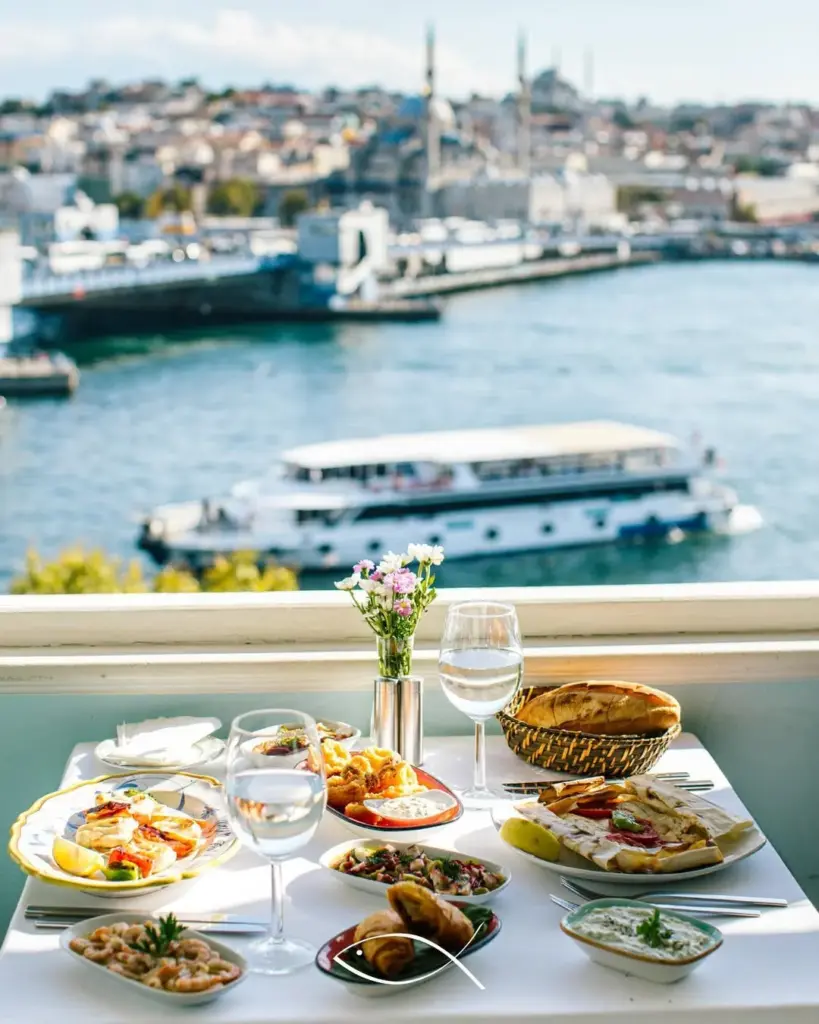 Masada taze deniz mahsulleri, çeşitli mezeler, ekmek ve su dolu kadehlerle birlikte, arka planda İstanbul'un ışıl ışıl denizi, hareketli tekneleri ve tarihi şehir silüeti görünüyor, bu görüntü Karaköy'deki bir balıkçıda keyifli bir yemeği betimliyor.