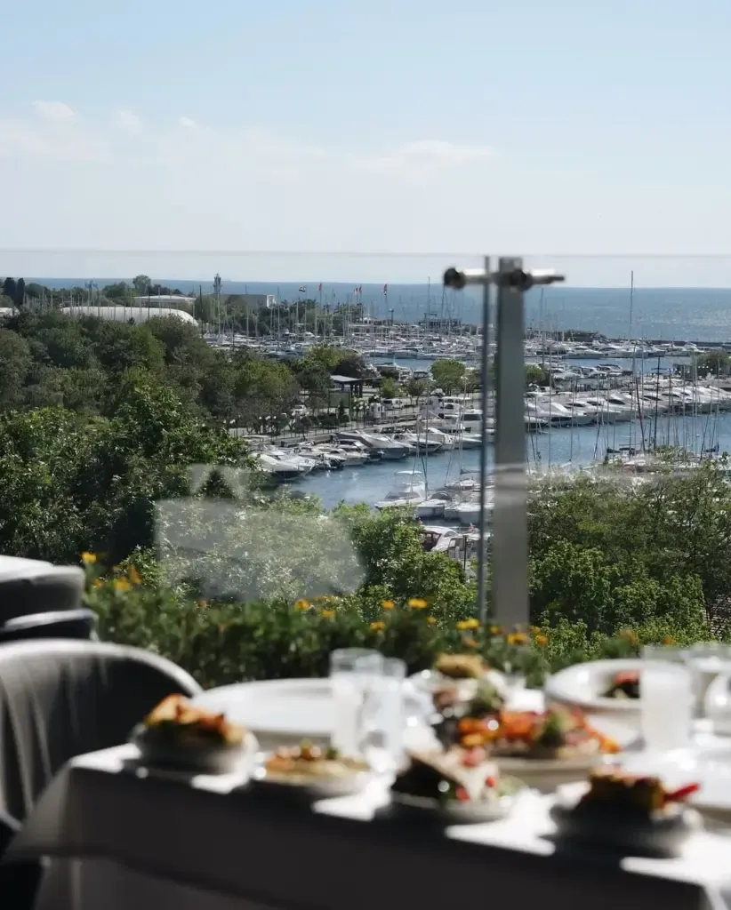 Bir masanın üzerinde bulanık duran çok sayıda meze tabağı ön plandayken, cam bir bariyerin ardında berrak mavi denize uzanan bir marinanın ve teknelerin panoramik manzarasının göründüğü güneşli bir restoran terası, ouzo keyfi için ideal bir ortam sunuyor.