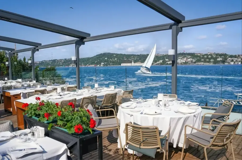 Açık hava Park Fora restoran terası, beyaz örtülü masa ve sandalyelerle dolu, ön planda kırmızı sardunyalar, arka planda Boğaz'ın mavi sularında süzülen beyaz yelkenli ve karşı kıyıdaki yeşil tepelerle beraber harika bir yemek deneyimi sunuyor.