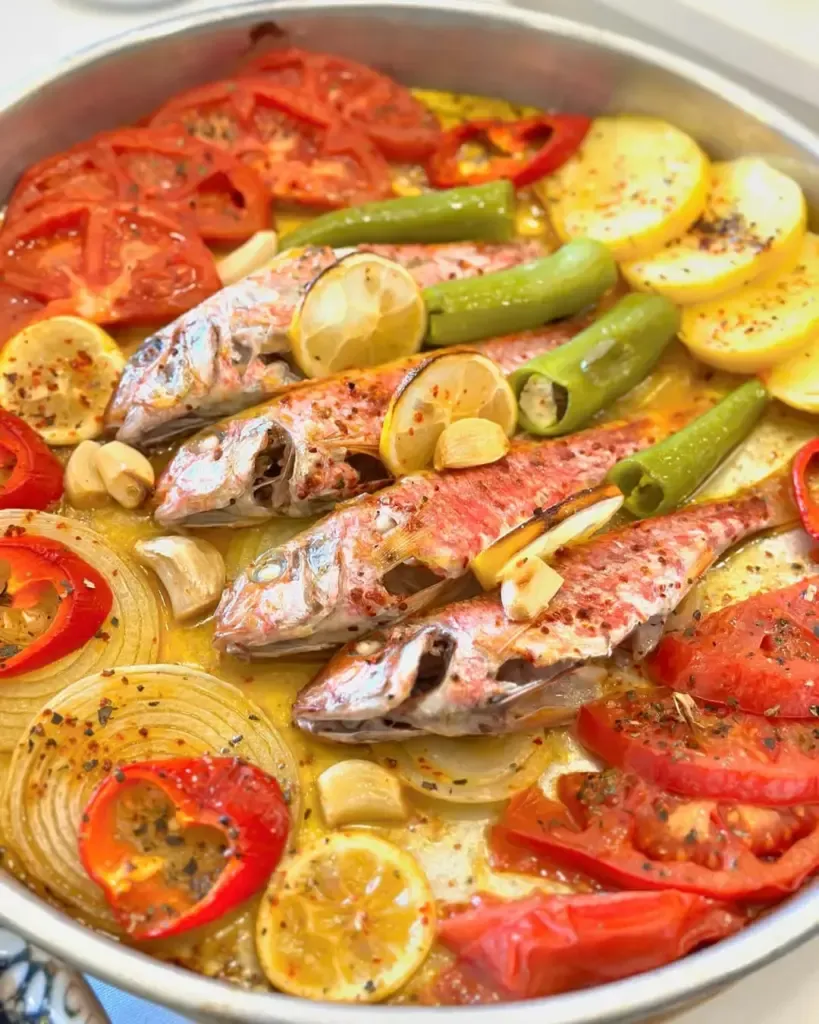 Büyük, parlak metal bir tepside, zeytinyağında pişmiş ve üzerine baharatlar serpilmiş, limon dilimleri ve sarımsak taneleri ile süslenmiş barbun balıkları; çevresine düzenli şekilde yerleştirilmiş taze domates, patates, halka soğan, yeşil ve kırmızı biber dilimleriyle görsel olarak zengin bir deniz mahsulü yemeği sunuluyor.